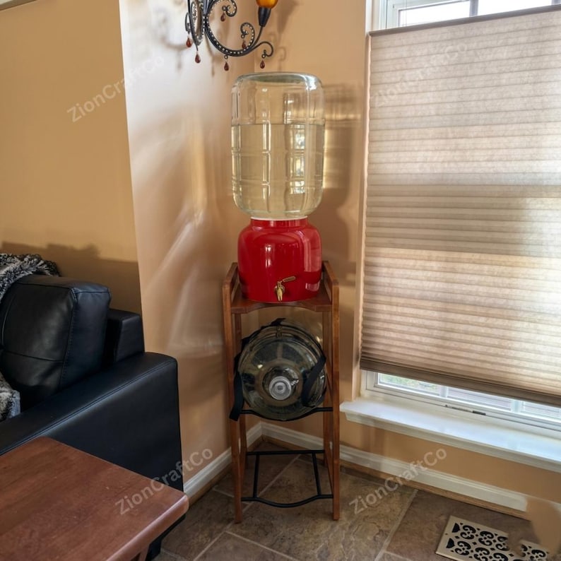 May include: A wooden water dispenser stand with a red ceramic base and a clear glass water jug on top. A second empty glass water jug with a black carrying strap is stored on the lower shelf of the stand. The stand has a tall, rectangular frame.