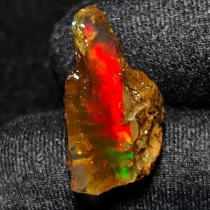 May include: A rough-cut opal gemstone with a vibrant display of color. The opal features a striking red core, transitioning to orange and yellow hues, with hints of green. The stone has a natural, earthy appearance with a textured, brown matrix.