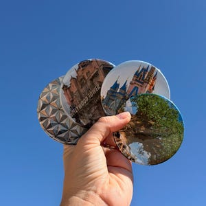 May include: A hand holding a collection of round coasters featuring iconic images. One coaster displays the Hollywood Tower Hotel, another shows a castle, and a third features a tree. The background is a clear blue sky.