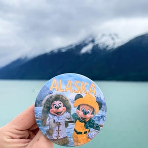 May include: A circular pinback button featuring a graphic of Minnie Mouse in a white fur-trimmed coat and Mickey Mouse in a yellow rain hat and overalls. The word ALASKA is printed in large orange letters across the top of the button design.