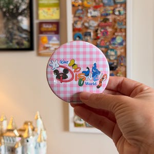 May include: A round pinback button featuring a pink and white gingham pattern background. The text Walt Disney World is printed in the center, decorated with various character-themed icons including a glove, ears, a hat, and a tail.