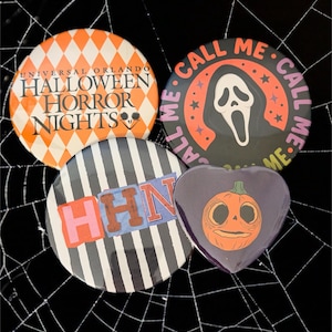 May include: Four circular and heart-shaped pinback buttons featuring Halloween themes. One button has an orange diamond pattern with text reading Universal Orlando Halloween Horror Nights. Another shows a ghost mask with the text Call Me. A third has black and white stripes with the letters HHN. The fourth is a heart-shaped button with a pumpkin illustration.