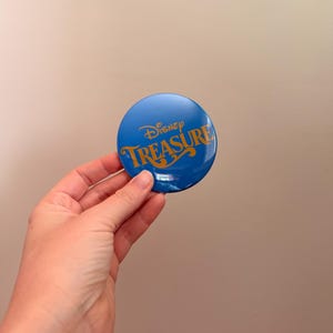 May include: A round, blue button with the word "Disney" above the word "Treasure" in gold script. The button is held in a hand against a neutral background. The button is a collectible item.