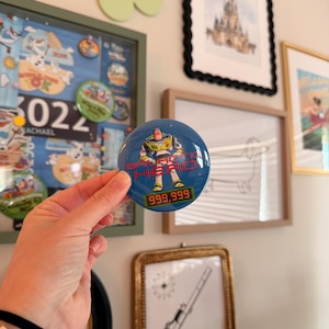 May include: A round, blue button featuring Buzz Lightyear from Toy Story. The button has the text "Buzz Hero" and a numerical display reading "999,999". The button is held in front of a wall with framed artwork and a decorative shelf.