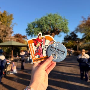 May include: A rectangular Walt Disney World annual passholder sticker featuring Nick Wilde and Judy Hopps, alongside a circular light blue button with the text here for my magnet and a small graphic of Mickey Mouse holding a magnet.
