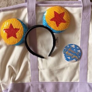 May include: A cream canvas tote bag with light purple straps and base, featuring a black headband with sequined yellow, blue, and red star ears, and a round blue pin with white clouds and the text I knew it, i knew you printed in gold.