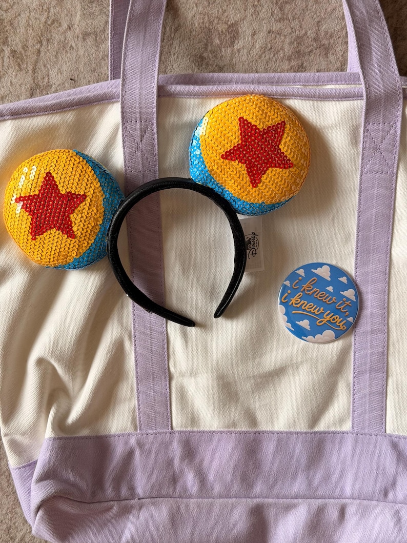 May include: A cream canvas tote bag with light purple straps and base, featuring a black headband with sequined yellow, blue, and red star ears, and a round blue pin with white clouds and the text I knew it, i knew you printed in gold.
