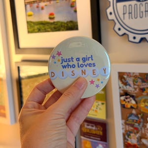 May include: A round, cream-colored pinback button featuring the blue text 'just a girl who loves' above the word 'DISNEY' in white letters on individual light blue circles. The design includes small pink, purple, and blue star accents.