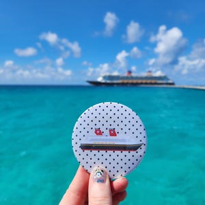 May include: A circular pinback button featuring a white background with a black polka dot pattern and a graphic illustration of a cruise ship with two red smokestacks, a black hull, and a yellow stripe along the side of the vessel.