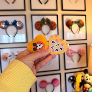 May include: Two heart-shaped buttons. One is orange with a Mickey Mouse graphic and text reading Walt Disney World Passholder. The other is yellow with pink polka dots and the black letters WDW AP arranged in a square grid pattern.