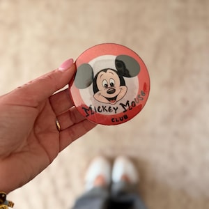 May include: A round, red and white Mickey Mouse Club button. The button features a smiling Mickey Mouse face with black eyes and a red mouth. The text "Mickey Mouse Club" is written in red.