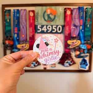 May include: A circular button featuring a pink party hat with colorful ribbons, a pink star wand, and small yellow stars on a white background. The text "just a Whimsy girlie" is printed in a cursive pink font across the center of the button.