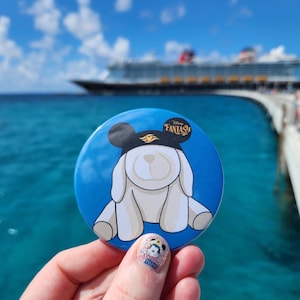 May include: A circular button featuring a white plush dog wearing black mouse ears with a gold emblem. The background is a solid blue, and the text Disney Fantasy is printed in the upper right corner of the button design in a dark font.
