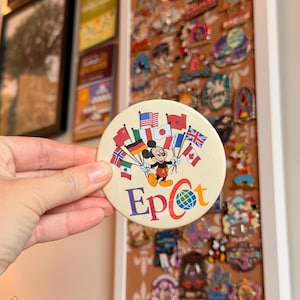 May include: A round, off-white button featuring a central illustration of Mickey Mouse holding a cluster of various international flags. Below the character, the word Epcot is printed in colorful, bold lettering with a globe icon replacing the letter o.