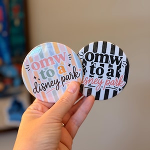 May include: Two circular pin-back buttons featuring the text "omw to a disney park". One button has vertical pastel stripes in pink, blue, and yellow. The other button features bold black and white vertical stripes with pink cursive text.