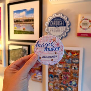 May include: A round, pink and white button with the text "magic maker" and a star-topped wand. Below the text is a name tag that reads "@ANGIETORRESWILSON" and "TRAVEL AGENT."
