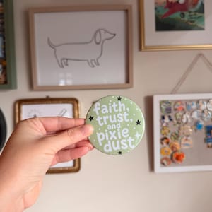 May include: A round, light green pin-back button featuring white text that reads faith, trust, and pixie dust. The design includes small white dots and black star icons scattered across the surface, creating a decorative graphic element.