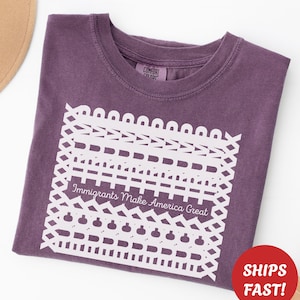 May include: A purple t-shirt with a white graphic design and the text "Immigrants Make America Great." The shirt is folded and has a round neck. A red circle with the words "SHIPS FAST!" is visible.