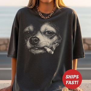 May include: Dark gray t-shirt featuring a detailed black and white illustration of a dog smoking a cigar. The dog's face is the focal point, with smoke rising from the cigar. The shirt has short sleeves and a relaxed fit.