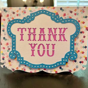 May include: A decorative thank you card with a white background covered in colorful confetti. The card features a blue frame with a white interior, and the words "THANK YOU" are displayed in pink, cut-out letters.