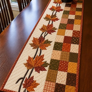 May include: A rectangular quilted table runner featuring a cream-colored section with an appliqued vine of multi-colored autumn maple leaves and a side panel composed of a patchwork grid of squares in shades of orange, brown, green, and cream.