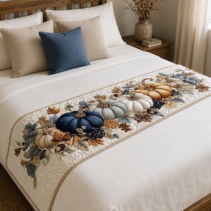 May include: A white bed runner featuring a quilted pattern with a central design of pumpkins in shades of blue, white, and orange, accented with brown leaves and berries. The runner is laid across a white bedspread with beige and blue throw pillows.