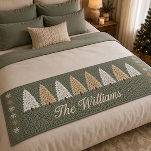 May include: A sage green quilted bed runner featuring an embroidered pattern of alternating white and tan Christmas trees, small snowflake accents, and the text "The Williams" in white script, laid across a neutral-colored bedspread.