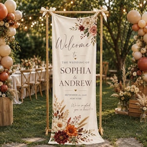 May include: A vertical cream-colored fabric wedding welcome sign suspended from a wooden frame. The text reads: Welcome to the wedding of Sophia & Andrew, September 10, 2026, New York. We're so glad you're here! Decorated with floral illustrations.