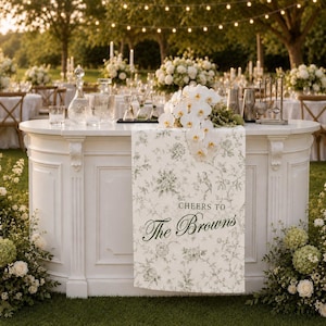 May include: A white outdoor bar with a floral runner that reads "Cheers to The Browns." The bar is decorated with glassware, bar tools, and a bouquet of white orchids. The background features tables, chairs, and string lights.