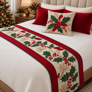 Op de afbeelding: Een bedloper en een vierkant sierkussen met een feestelijk hulst- en bessenpatroon op een crèmekleurige achtergrond met donkerrode fluwelen randen. De set ligt op een witte doorgestikte sprei met effen rode fluwelen kussens.