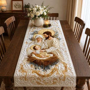 May include: A rectangular table runner featuring a quilted design of the Holy Family. The central image depicts a man, a woman, and an infant in a manger, all with gold halos. The fabric is white with gold and tan scrollwork and floral patterns.