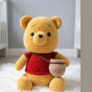 May include: A handmade, yellow crochet bear wearing a red shirt and carrying a small brown honey pot. The bear has black eyes and a black nose. The plush toy is sitting on a white, fluffy surface.