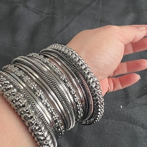 May include: A set of stacked silver-toned metal bangles featuring a variety of textures, including smooth polished bands, intricate beaded edges, and delicate floral-patterned embossed designs, worn together as a cohesive wrist accessory.