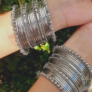 Op de afbeelding: Twee polsen versierd met meerdere zilveren armbanden. Elke armband heeft een gepolijste zilveren kleur, met een kralenontwerp langs de randen. De armbanden zijn gestapeld, waardoor een gelaagd effect ontstaat. De achtergrond is een wazig groen, wat op gebladerte duidt.