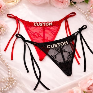 Puede incluir: Dos prendas interiores tipo tanga, una roja y otra negra, con la palabra "CUSTOM" escrita con pedrería. El tanga rojo tiene lazos rojos, y el tanga negro tiene lazos negros. Ambos son de encaje.