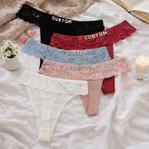 Puede incluir: Cinco tangas en negro, rojo, azul claro, rosa y blanco. Cada uno tiene un borde de encaje y la palabra "CUSTOM" en plata. La ropa interior está dispuesta sobre una superficie blanca. La imagen incluye una vela y flores.