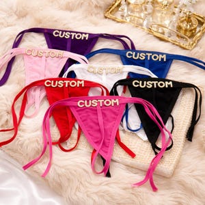 Puede incluir: Una colección de tangas personalizados en varios colores, incluyendo morado, rosa, rojo, blanco, azul y negro. Cada tanga presenta la palabra "CUSTOM" en letras brillantes. La ropa interior tiene lazos laterales.