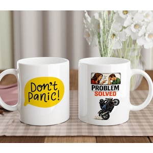 May include: Two white ceramic mugs. One mug features the text "Don't Panic!" in a yellow speech bubble. The other mug has the text "Problem Solved" with a motorcycle wheelie illustration.