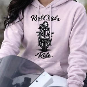 May include: A light pink hoodie with the text "Real Chicks Ride" and a graphic of a woman on a motorcycle. The hoodie is worn by a person sitting on a white motorcycle.