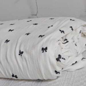 May include: A folded white fabric featuring a repeating pattern of small, dark black bows. The material has a subtle vertical ribbed texture throughout, creating a soft and layered appearance with the bow print evenly distributed across the surface.
