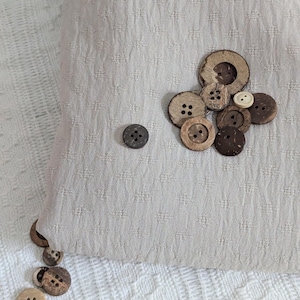 May include: A collection of various sized wooden buttons, some with two and four holes, scattered on a light beige textured fabric. The buttons are in shades of brown and tan, with a few lighter-colored buttons interspersed.