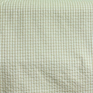 Op de afbeelding: Een stuk stof met een klassiek beige en wit gingham ruitpatroon. Het materiaal heeft een uitgesproken seersucker textuur met een geplooid oppervlak, waardoor een raster van kleine vierkantjes over het gehele textiel ontstaat.