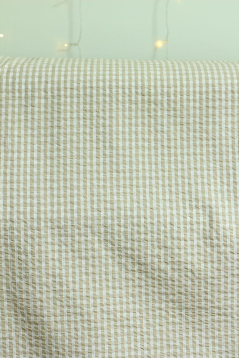 Op de afbeelding: Een stuk stof met een klassiek beige en wit gingham ruitpatroon. Het materiaal heeft een uitgesproken seersucker textuur met een geplooid oppervlak, waardoor een raster van kleine vierkantjes over het gehele textiel ontstaat.
