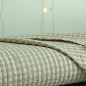 Op de afbeelding: Een gevouwen textiel met een lichtbeige en wit gingham ruitpatroon. De stof heeft een zichtbare gekreukelde textuur, waarbij de vouwen gelaagde secties cre&euml;ren die het consistente ruitpatroon over het gehele oppervlak laten zien.