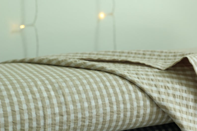 Op de afbeelding: Een gevouwen textiel met een lichtbeige en wit gingham ruitpatroon. De stof heeft een zichtbare gekreukelde textuur, waarbij de vouwen gelaagde secties cre&euml;ren die het consistente ruitpatroon over het gehele oppervlak laten zien.