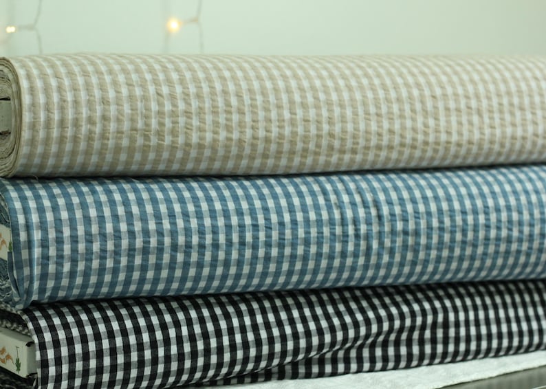 Op de afbeelding: Drie horizontaal gestapelde rollen stof, elk met een klassiek gingham ruitpatroon. De bovenste rol is beige en wit, de middelste rol is blauw en wit en de onderste rol is zwart en wit. De stof heeft een zichtbaar textuurrijk oppervlak.