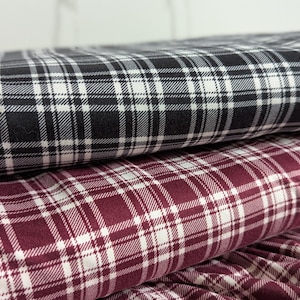 May include: Close-up of two rolls of plaid fabric. The top roll features a black and white pattern, while the bottom roll displays a burgundy and white design. The fabric is tightly woven, with a classic plaid design.