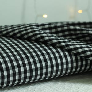 Op de afbeelding: Een gevouwen stuk stof met een klassiek zwart-wit vichy-ruitpatroon. Het textiel vertoont een consistent raster van kleine, afwisselende vierkantjes over het oppervlak, met een zachte textuur en zachte plooien en gedrapeerde randen.
