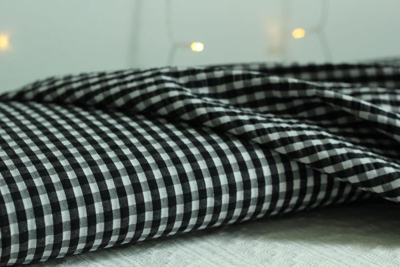 Op de afbeelding: Een gevouwen stuk stof met een klassiek zwart-wit vichy-ruitpatroon. Het textiel vertoont een consistent raster van kleine, afwisselende vierkantjes over het oppervlak, met een zachte textuur en zachte plooien en gedrapeerde randen.