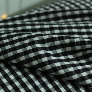 Op de afbeelding: Een stuk stof met een zwart-wit gingham ruitpatroon. Het materiaal heeft een uitgesproken gekreukelde, bobbelige textuur over het gehele oppervlak, wat zorgt voor zichtbare plooien en diepte in het geruite textielmateriaal.
